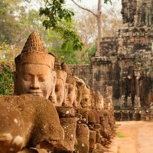 Una fila di statue di pietra erose dal tempo, con volti scolpiti, fiancheggia un sentiero sterrato che porta a un antico portale in pietra.