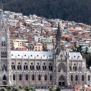 Une grande cathédrale gothique grise avec un clocher se dresse devant une ville dense bâtie sur une colline boisée.