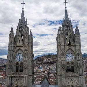 Dos torres de reloj de catedral de estilo gótico dominan una ciudad extensa con colinas al fondo bajo un cielo nublado.