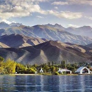 Un lago en calma se extiende frente a una orilla con árboles verdes y edificios blancos, con una vasta y ondulante cordillera de fondo bajo un cielo parcialmente nublado.