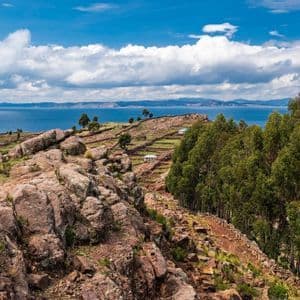Una ladera rocosa con campos verdes aterrazados y árboles domina un gran lago azul bajo un cielo parcialmente nublado.