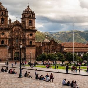 An ornate stone cathedral with twin bell towers overlooks a busy, paved city square filled with people, with green mountains in the background.