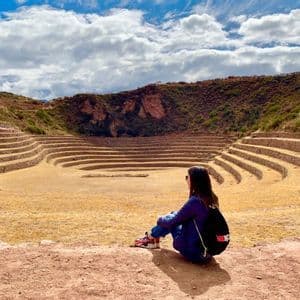 Una donna con uno zaino siede su una sporgenza sterrata che si affaccia su un grande sito archeologico terrazzato e circolare sotto un cielo nuvoloso.