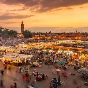 An aerial view of a bustling city market square filled with illuminated stalls and crowds of people at sunset.