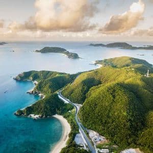 Una vista aerea di isole lussureggianti e verdi, con una strada costiera tortuosa che si snoda accanto a acque cristalline e turchesi sotto un cielo parzialmente nuvoloso.