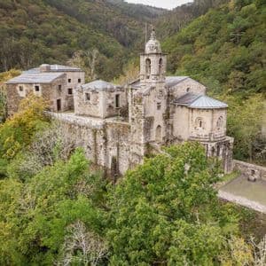 Una vista aérea de un antiguo monasterio de piedra con un campanario, enclavado en una ladera cubierta de densos árboles verdes.