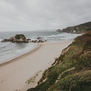 Una vista desde un acantilado cubierto de hierba con vistas a una playa de arena con olas y afloramientos rocosos bajo un cielo nublado.
