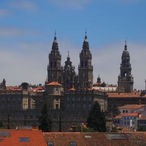 Una ornamentada catedral de piedra con múltiples agujas se eleva sobre los tejados de tejas rojas de una ciudad contra un cielo azul.
