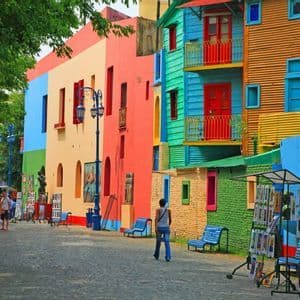 Una calle adoquinada, bañada por el sol, está bordeada de edificios vibrantes y multicolores mientras la gente camina y explora puestos de arte al aire libre.