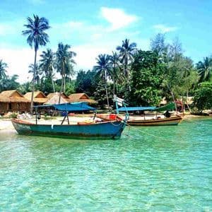 Due barche sono ormeggiate in acque limpide e turchesi, vicino a una spiaggia sabbiosa con capanne di paglia e palme.