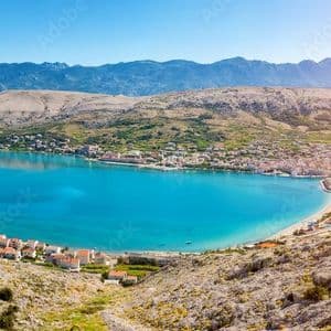 Panoramica di una cittadina costiera con case dai tetti rossi lungo una vivace baia blu, circondata da colline rocciose e montagne lontane.