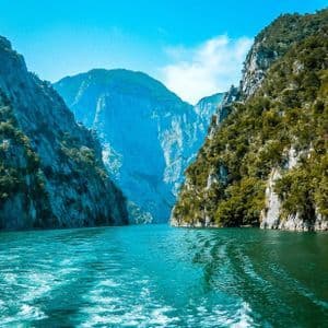 View from a boat on a turquoise river winding through a steep, forested mountain canyon under a blue sky.