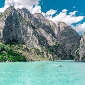 A small boat moves across turquoise water in a canyon surrounded by steep, rocky mountains with green slopes under a blue sky with white clouds.