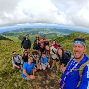 Un grupo de viaje de WeRoad se toma una selfie en una colina cubierta de hierba con vistas a un lago de cráter y al océano bajo un cielo nublado.