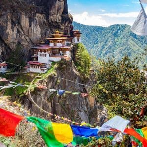 Un monastero bianco e rosso è incastonato in una scogliera a strapiombo, con bandiere di preghiera colorate in primo piano e montagne boscose sullo sfondo.