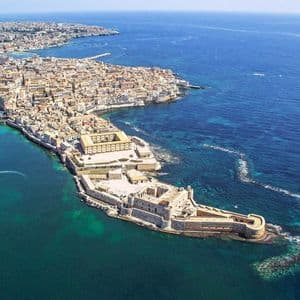 Una vista aerea di una storica città costiera e fortezza su una penisola, circondata da mare turchese e blu intenso.