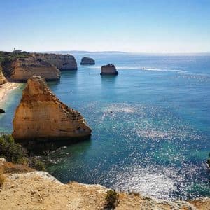 Un capo gruppo WeRoad con un cappello siede su una scogliera che si affaccia su una costa rocciosa con archi naturali sul mare e una piccola spiaggia.
