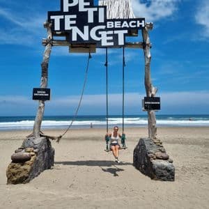 Eine Frau sitzt auf einer großen Holzschaukel an einem Sandstrand unter einem Schild mit der Aufschrift 'Petitenget Beach', mit dem Meer im Hintergrund.