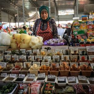 Una donna che indossa un foulard e un grembiule si trova dietro una vivace bancarella del mercato che vende spezie, noodles e altri prodotti alimentari.
