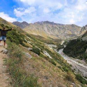 Un uomo con le braccia distese sta su un sentiero di montagna che domina una vasta valle con cime rocciose sotto un cielo parzialmente nuvoloso.