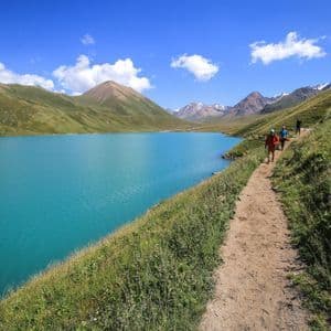 Un viaggio di gruppo WeRoad in trekking su un sentiero sterrato, accanto a un lago alpino turchese, circondato da montagne verdi sotto un cielo azzurro limpido.