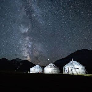 Tre yurte illuminate riposano in un campo ai piedi delle montagne sotto un cielo stellato con la Via Lattea.