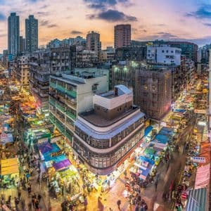 Una vista grandangolare dall'alto di un vivace mercato cittadino al tramonto, con folle di persone tra bancarelle illuminate e alti edifici.