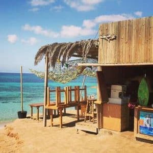 A rustic wooden beach bar with a thatched roof stands on a sandy shore, overlooking a calm turquoise sea under a blue sky.