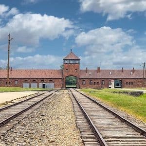 Dos conjuntos de vías de tren conducen hacia la entrada de un gran edificio de ladrillo con una torre de vigilancia central bajo un cielo azul con nubes.