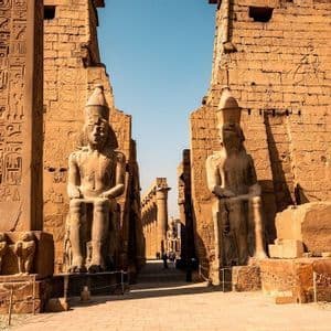 Große Steinstatuen von Pharaonen flankieren einen sonnendurchfluteten Korridor, der zu einem altägyptischen Tempel führt, dessen hohe Mauern mit Hieroglyphen bedeckt sind.