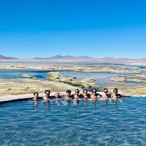 Un groupe WeRoad en voyage prend la pose dans une source chaude naturelle, avec un vaste paysage aride en arrière-plan.
