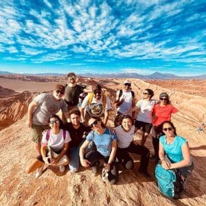 A WeRoad group trip poses for a photo on a rocky peak overlooking a desert landscape under a bright blue, cloudy sky.