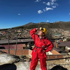 Une femme vêtue d'une combinaison de protection rouge et d'un casque jaune avec une lampe frontale se tient sur une colline surplombant une ville minière.