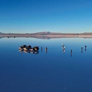 A WeRoad group trip stands on a vast, reflective salt flat with 4x4 vehicles, creating a perfect mirror of the blue sky and mountains.