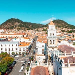 An elevated view of a cityscape with a white bell tower, orange tiled roofs, and green hills under a clear blue sky.