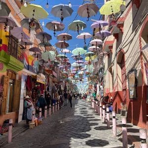 Colorful umbrellas hang over a busy cobblestone market street, casting distinct shadows on the ground as people walk between shops.