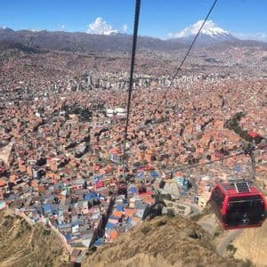 Un téléphérique rouge survole une ville tentaculaire dans une vallée, avec des montagnes enneigées sous un ciel bleu clair.