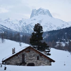 Una casa rustica in pietra si trova in una valle innevata, con un alto pino e montagne innevate sullo sfondo.
