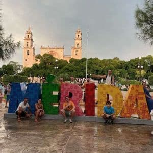 Un groupe WeRoad en voyage pose pour une photo devant de grandes lettres colorées formant 'MERIDA' sur une place avec une cathédrale en arrière-plan.
