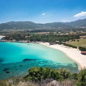 Vista aérea de una playa de arena que se curva por la costa, con aguas turquesas cristalinas y colinas verdes bajo un cielo azul.