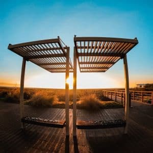 Dos bancos modernos con pérgolas de lamas en un paseo de madera, frente a un paisaje herboso durante un atardecer dorado bajo un cielo despejado.