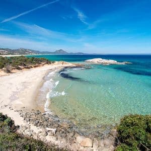 Una vista panoramica di una spiaggia isolata di sabbia bianca con acqua cristallina turchese, incorniciata da vegetazione verde sotto un vasto cielo blu.