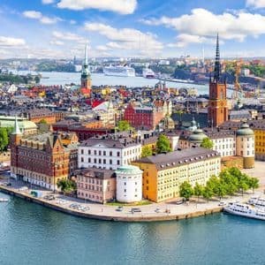 Vista aérea de una ciudad europea histórica con edificios coloridos y agujas a lo largo de un paseo marítimo bajo un cielo azul.
