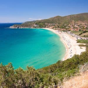 Una vista aérea de una playa de arena concurrida y en forma de media luna con agua turquesa, un pueblo costero y colinas verdes bajo un cielo azul claro.