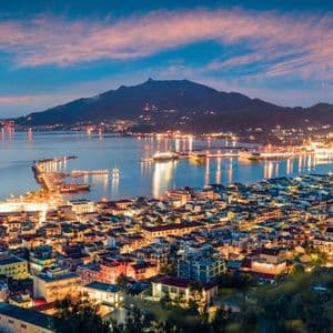 Una vista panoramica di una città costiera illuminata e del suo porto al tramonto, con una grande montagna stagliata contro un cielo colorato.