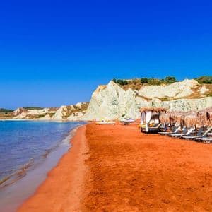 Una lunga spiaggia con sabbia rossa caratteristica, acque azzurre e bianche scogliere, attrezzata con lettini e ombrelloni di paglia.