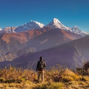 Un escursionista con uno zaino si trova su un crinale erboso, osservando una vasta catena di montagne innevate sotto un cielo azzurro e limpido.