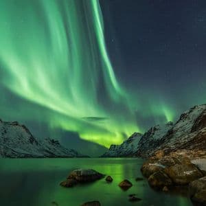 Corrientes verdes de la Aurora Boreal iluminan un cielo nocturno estrellado sobre un fiordo y montañas cubiertas de nieve.
