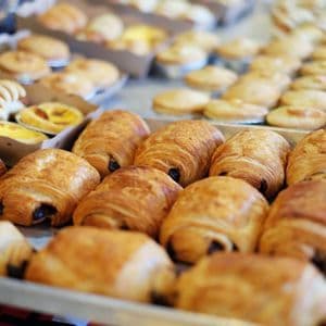 Tabletts mit frisch gebackenem Gebäck, mit goldbraunen Schokoladen-Croissants im Vordergrund einer Bäckereiauslage.