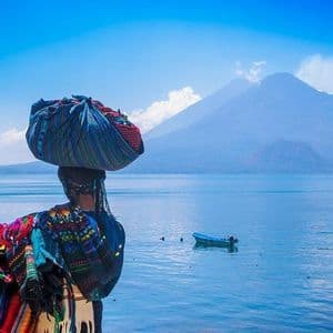 A person in colorful traditional clothing carries a bundle on their head, standing by a lake with large mountains in the background.